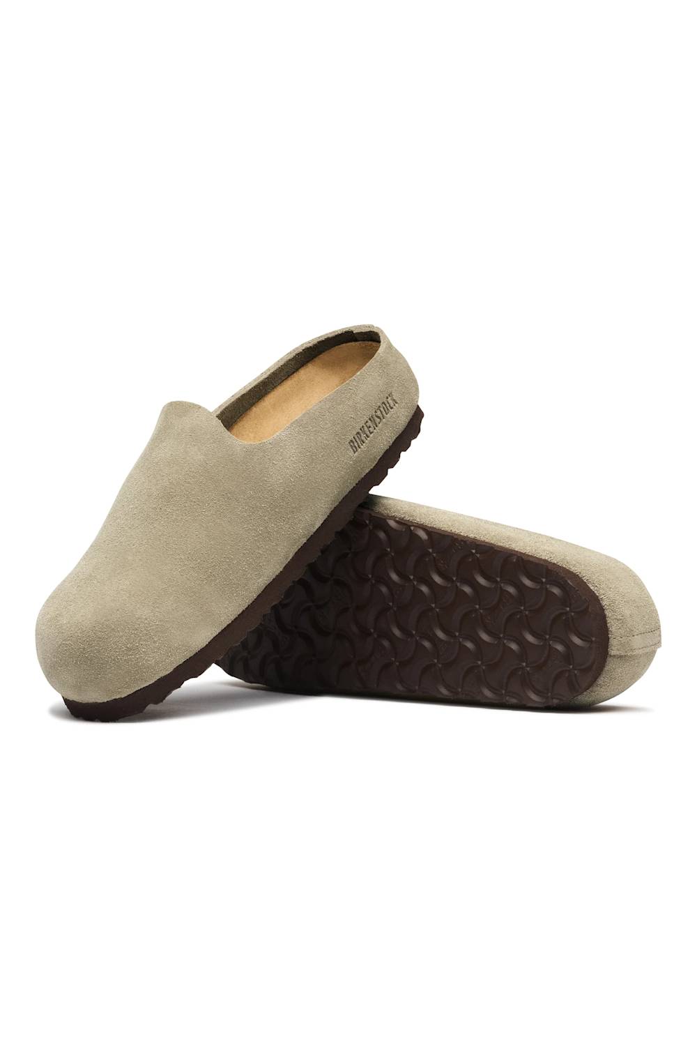A pair of slip-on clog-style slippers in light beige suede, with one shoe standing upright and the other lying on its side to show the dark brown textured sole. 