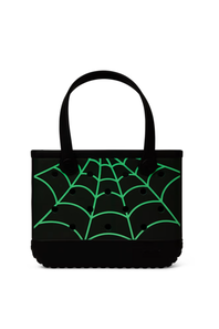 A black tote bag with a bright green glow in the dark spiderweb pattern printed across the front, featuring two black shoulder straps, set against a plain white background.