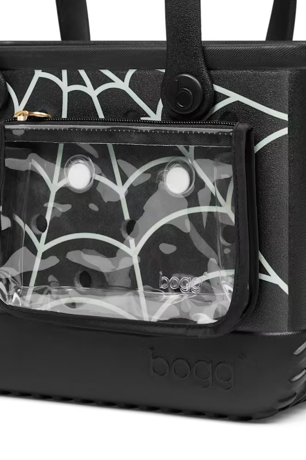 Close-up of a black Bogg tote bag with a clear front zip pocket showing a black-and-white camouflage patterned insert, plus a black handle and embossed Bogg branding on the base.
