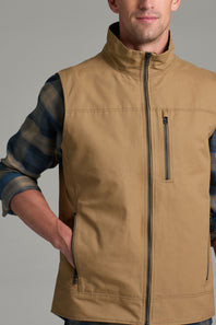 A man wearing a tan zip-front vest over a blue and brown plaid shirt, shown from the neck down against a plain gray studio background.