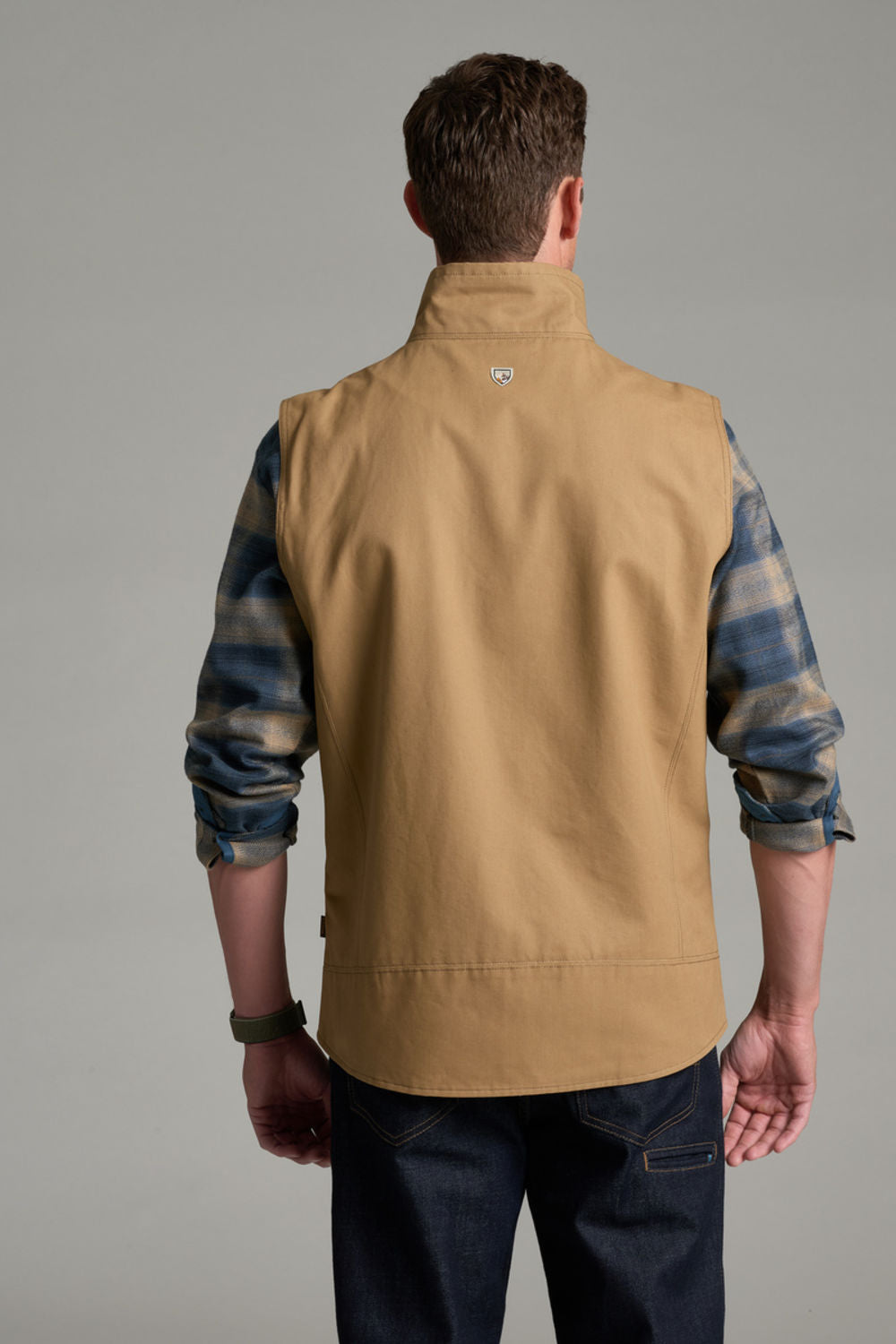 A man viewed from the back wearing a tan utility vest over a blue and gray plaid flannel shirt and dark jeans, against a solid gray studio background.