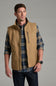 A man wearing a tan zip-front vest over a blue and tan plaid flannel shirt, paired with dark jeans, photographed against a plain gray studio background.