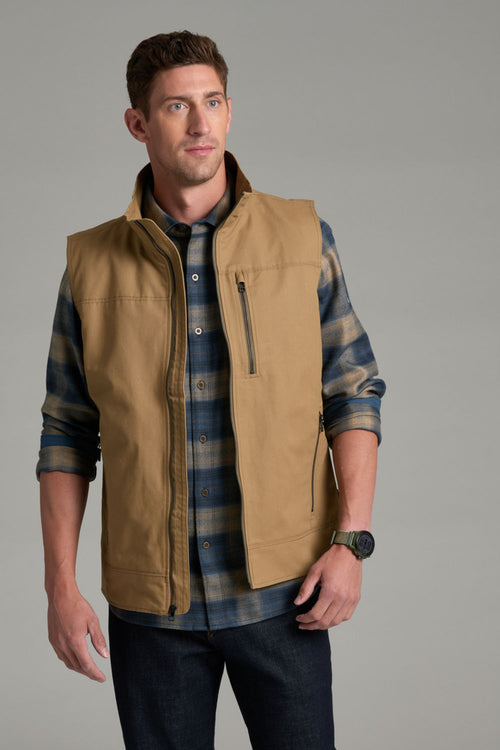 A man wearing a tan zip-front vest over a blue and tan plaid flannel shirt, paired with dark jeans, photographed against a plain gray studio background.