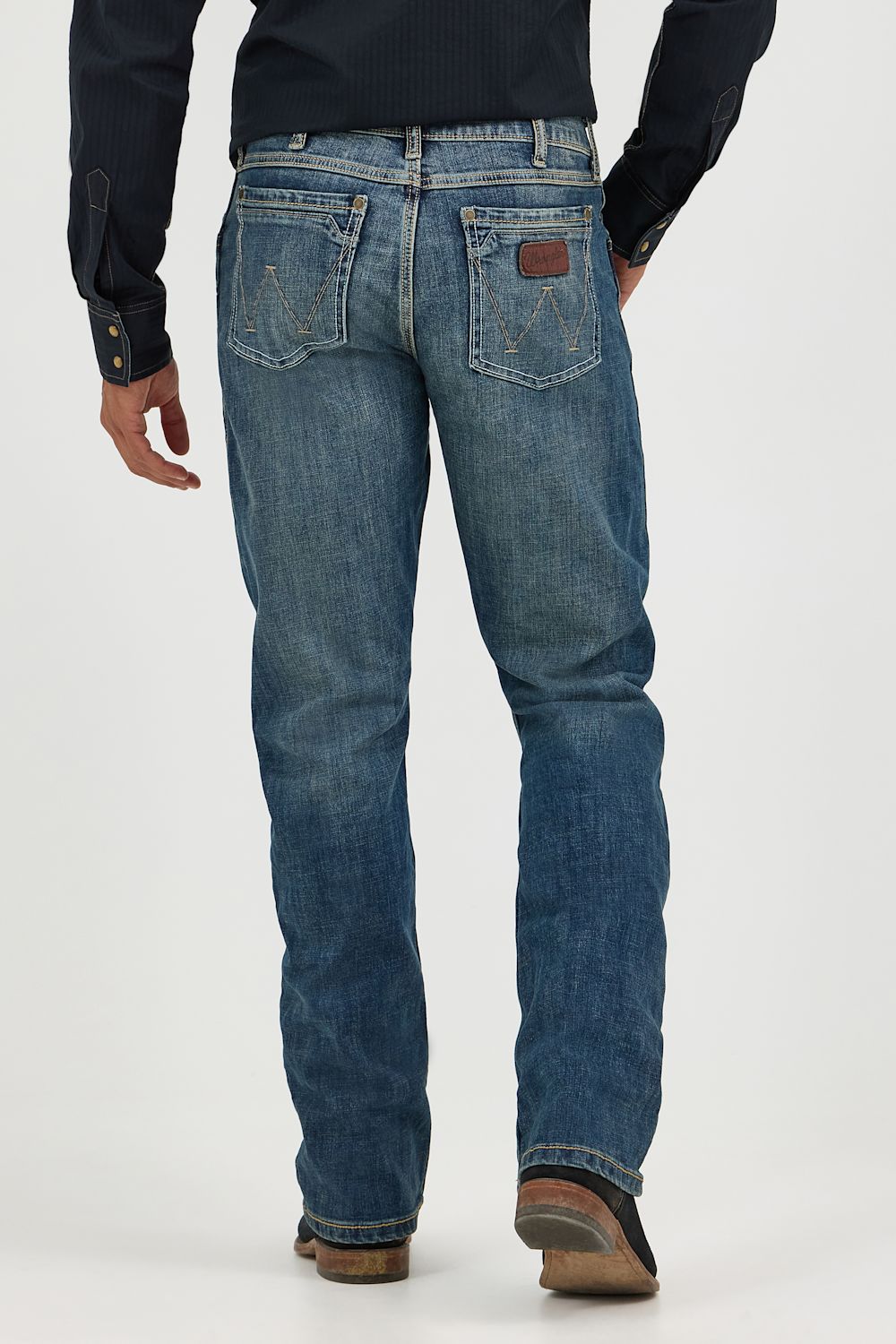 Rear view of a person wearing blue denim jeans with a dark navy shirt and brown shoes, standing against a light gray studio background.