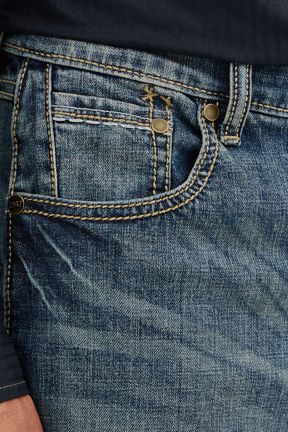 Close-up of the back pocket of blue denim jeans with contrast tan stitching, brass-tone rivets, and a small embroidered logo near the pocket.