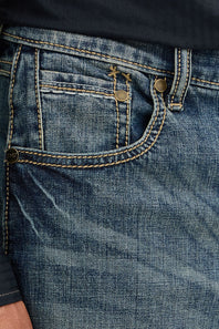 Close-up of the back pocket of blue denim jeans with contrast tan stitching, brass-tone rivets, and a small embroidered logo near the pocket.