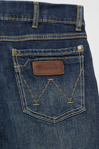 Close-up of the back pocket of blue denim jeans with contrast tan stitching, a brown leather Wrangler patch, and metal rivets on a light gray background.