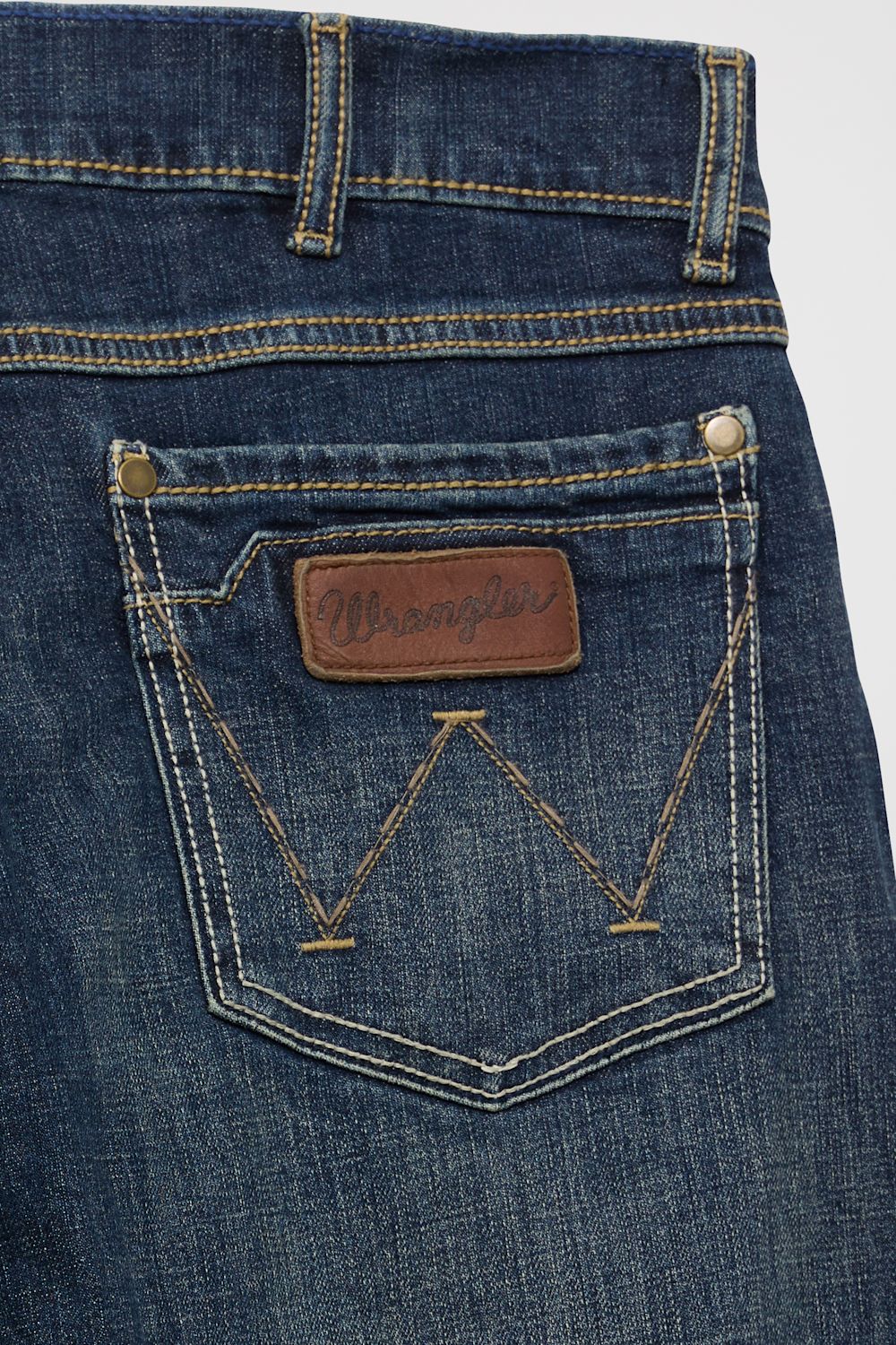 Close-up of the back pocket of blue denim jeans with contrast tan stitching, a brown leather Wrangler patch, and metal rivets on a light gray background.