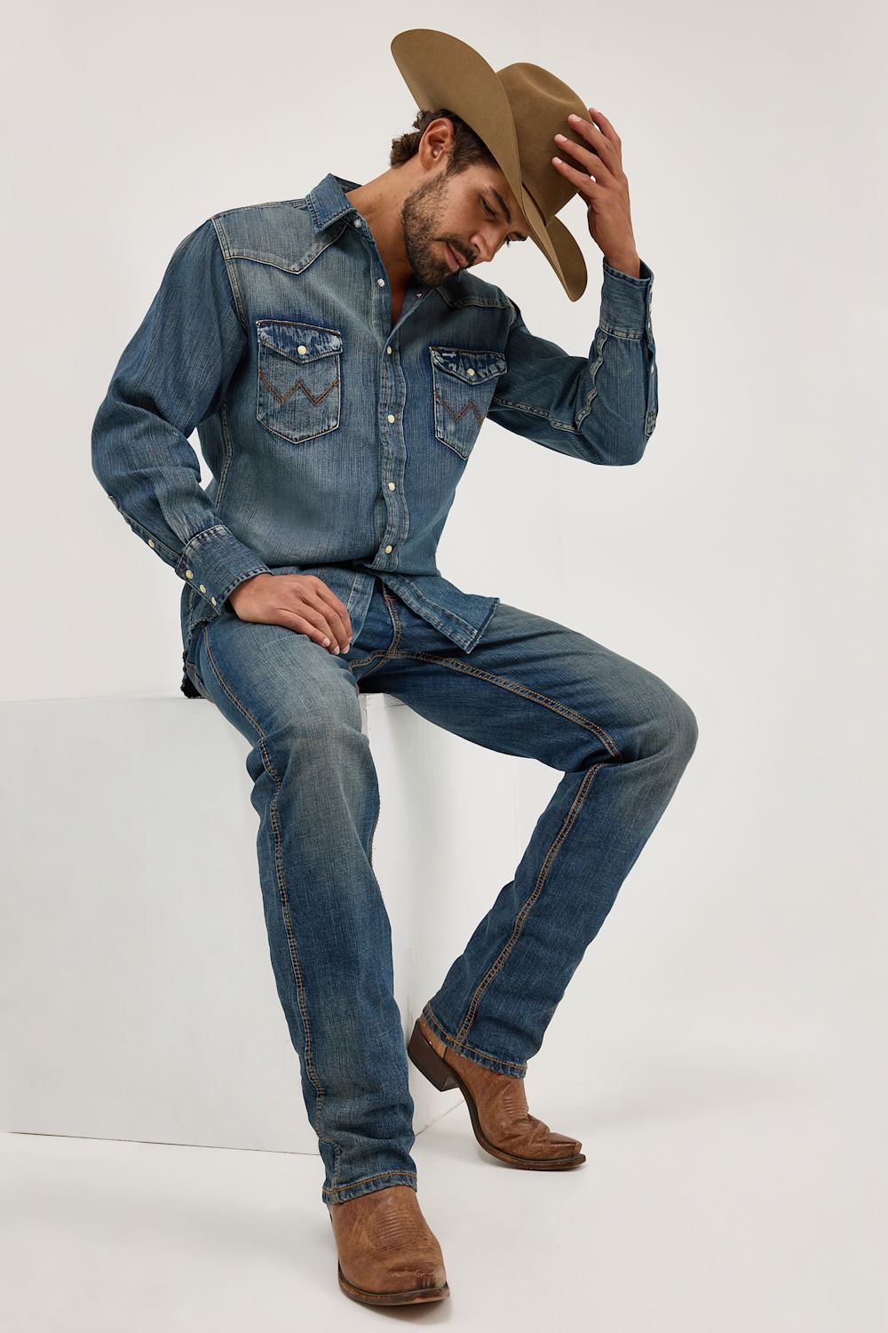 A man wearing a blue denim western shirt and matching jeans, sitting on a white block against a light gray studio background. He is also wearing a tan cowboy hat and brown boots.