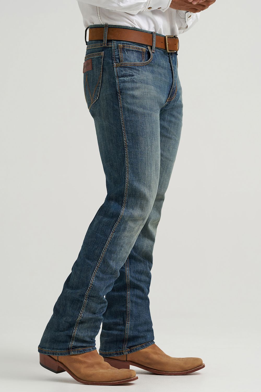 A person wearing blue denim jeans with a brown leather belt, paired with a white shirt and tan boots, shown from the waist down against a light gray studio background.