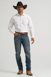 A man modeling a white long-sleeve western button-up shirt, blue jeans, a brown belt, brown cowboy boots, and a dark brown cowboy hat against a light gray studio background.