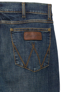 Close-up of the back waistband and pocket area of blue denim jeans, showing a brown leather Wrangler patch on the back pocket with gold-toned metal rivets and contrast stitching.