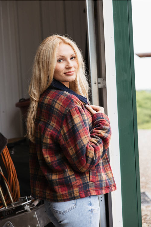 A woman with long blonde hair viewed from the back-side angle, wearing a burgundy, navy, and orange plaid barn jacket with a corduroy collar and light-wash denim jeans while looking back over her shoulder in a doorway.