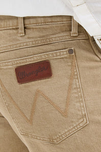 Close-up of the back pocket of Wrangler jeans in a light tan or khaki wash, showing the brown leather brand patch and contrast stitching.