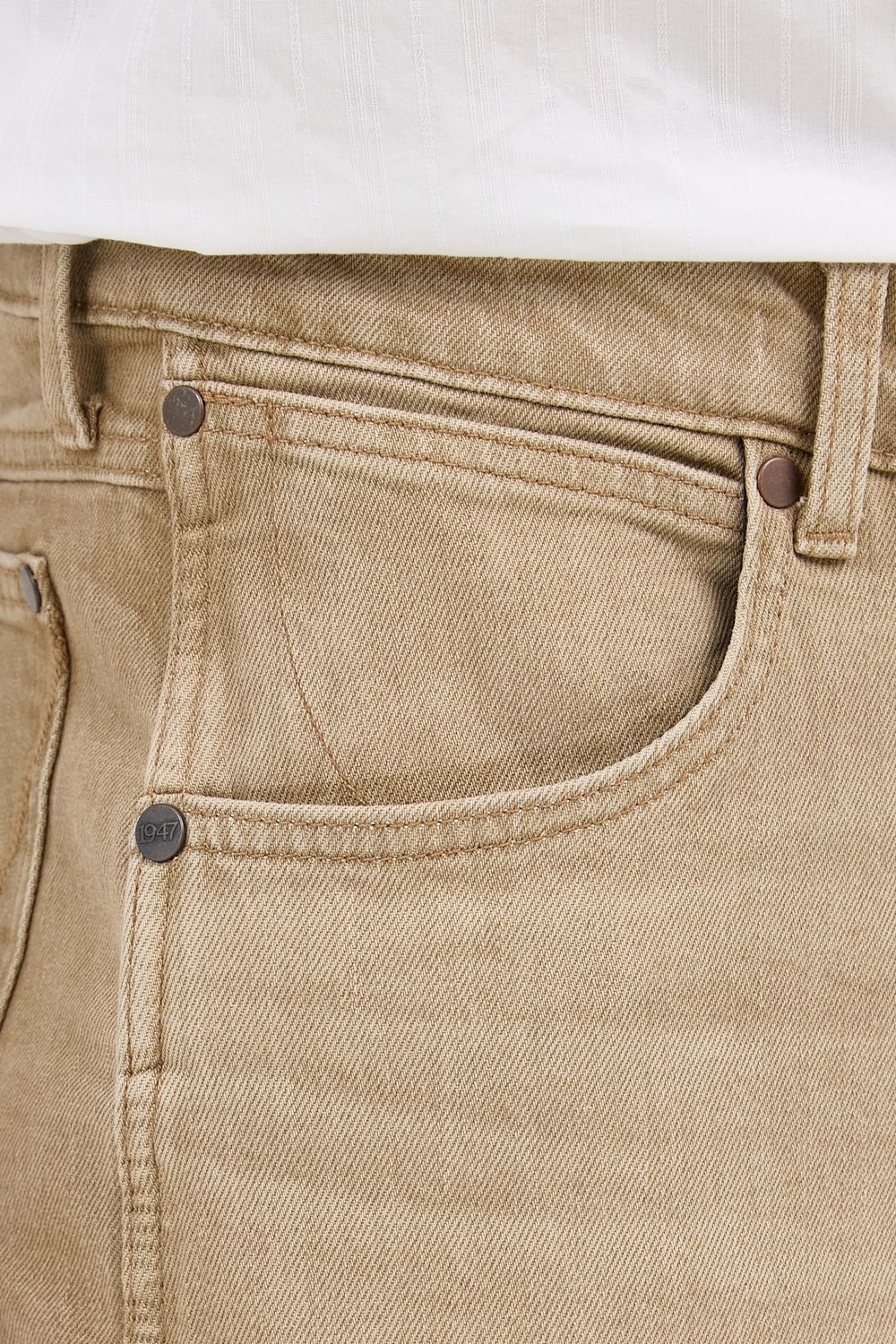 Close-up of beige denim jeans showing the front pocket, button, rivet, waistband, and a small portion of a white shirt at the top.