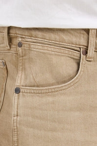 Close-up of beige denim jeans showing the front pocket, button, rivet, waistband, and a small portion of a white shirt at the top.