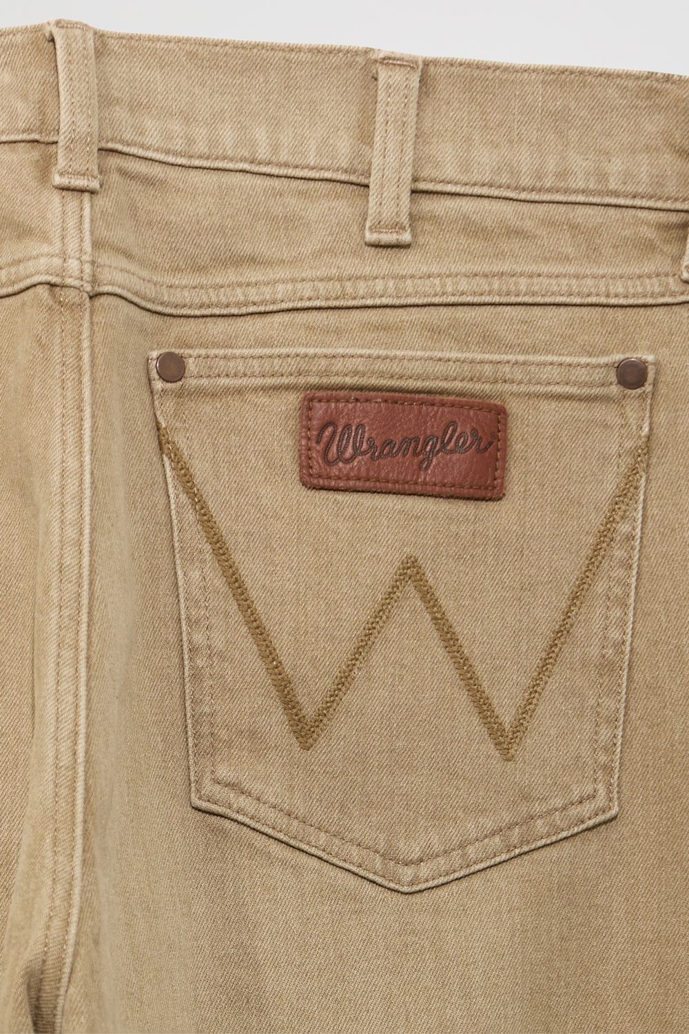 Close-up of the back pocket of tan denim jeans with a brown Wrangler brand patch and decorative stitching.