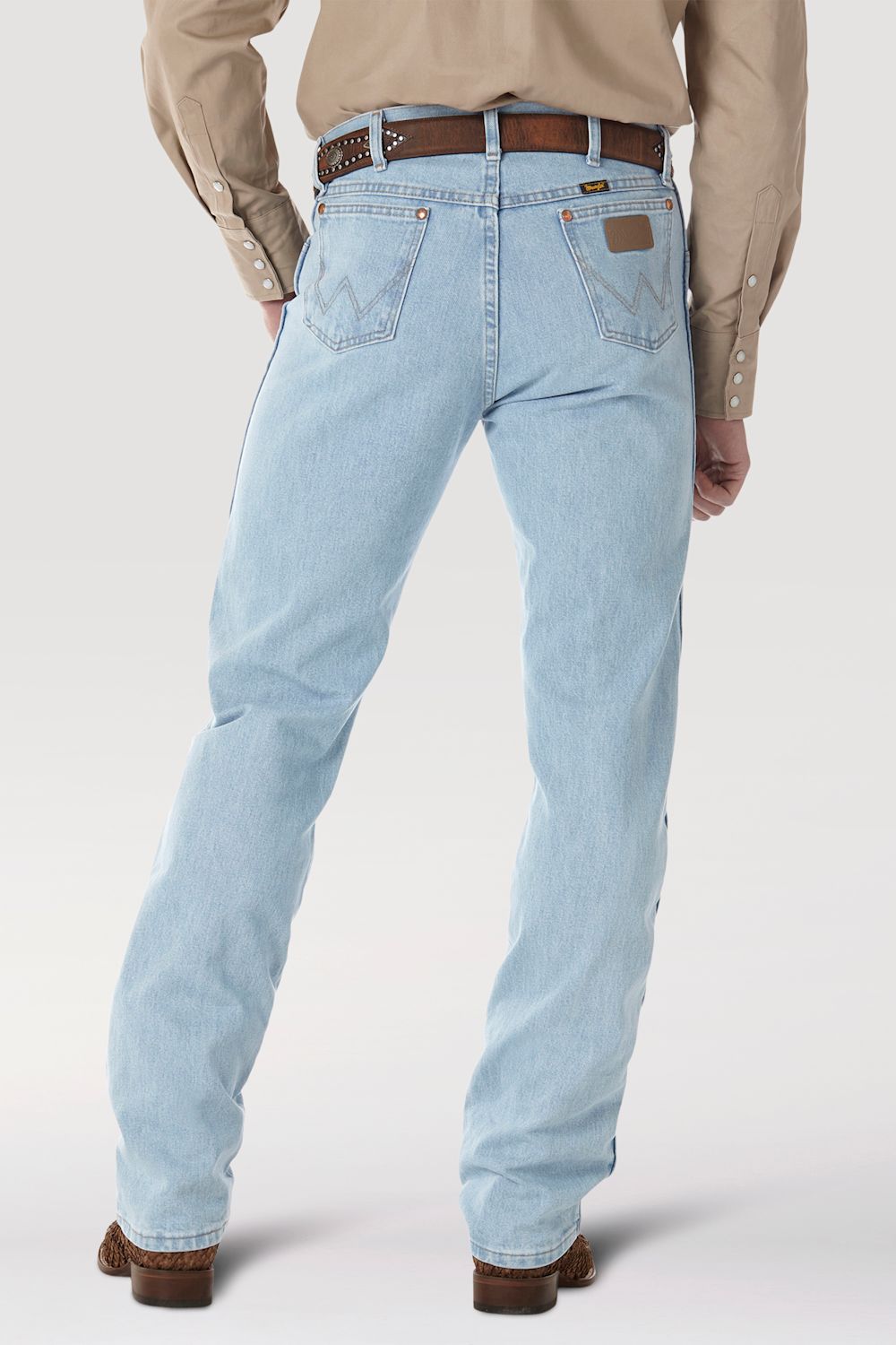 Rear view of a person wearing light-wash blue jeans with a brown belt, tan shirt, and brown shoes against a light gray studio background.