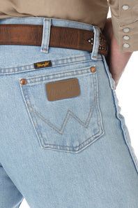 Close-up of the back waistband of light-wash blue denim jeans worn by a model, featuring a brown leather belt, a brown leather patch with white zigzag stitching, and a small black Wrangler brand label.