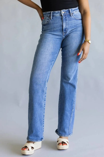 Woman shown from the waist down wearing medium wash denim jeans with a black top, gold watch and beige wedges standing in front of a plain studio background
