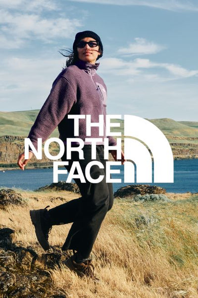 Person wearing a purple jacket with black pants walking in a grassy area with hills and  a lake in the background. Over top the image is THE NORTH FACE brand logo in white