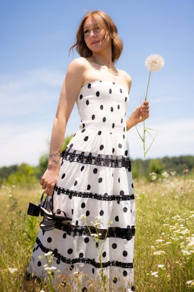 Woman standing and a grassy field holding a large dandelion and wearing a strapless white midi dress with black polka dots and holding black heels in the other hand