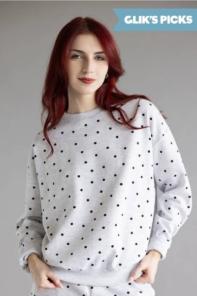 A woman wearing a grey crewneck sweatshirt with black polka dots and matching bottoms standing against a grey background and a blue flag badge in the upper right corner that read GLIK'S PICKS