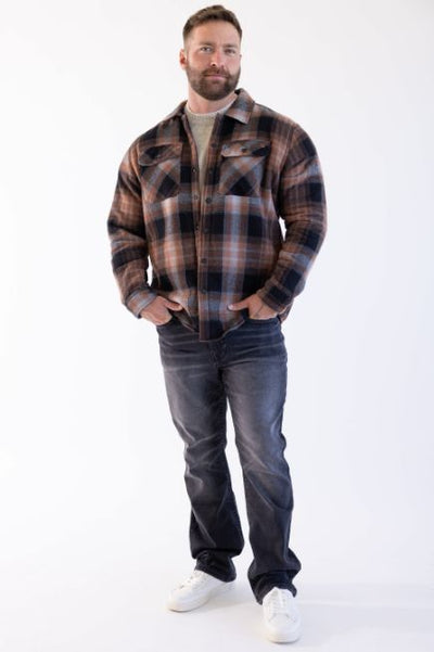 A man wearing a brown and beige colored flannel with dark wash denim jeans paired with a beige undershirt and white sneakers standing in front of a white background