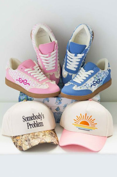 Display of colorful accessories featuring pink and blue lace-detail sneakers on stacked floral boxes, with two trucker hats in front—one reading 