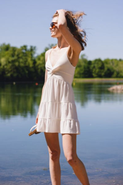 Girl standing in front of a lake wearing a short white dress and brown sunglasses
