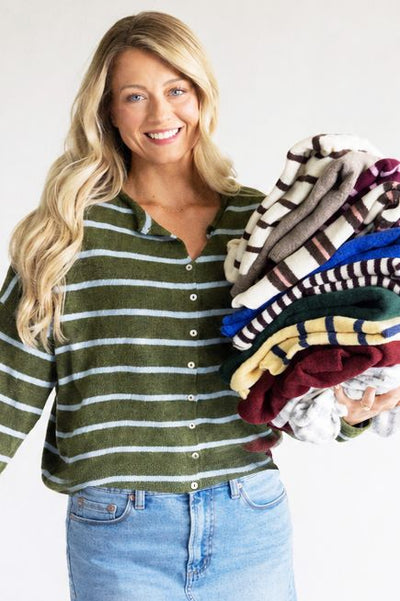 Woman wearing a dark green with blue stripes cardigan and medium wash blue denim jeans holding multiple different color and prints of sweaters standing against a plain background