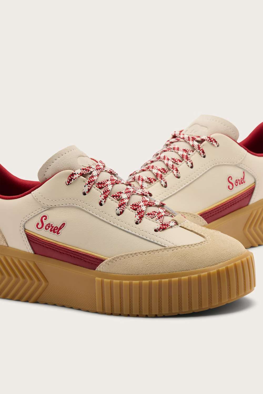 A pair of Sorel low-top sneakers in cream suede and leather, featuring red-and-white patterned laces, a red side stripe with the Sorel logo, and a tan gum sole.
