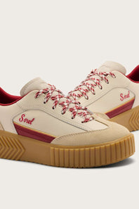 A pair of Sorel low-top sneakers in cream suede and leather, featuring red-and-white patterned laces, a red side stripe with the Sorel logo, and a tan gum sole.