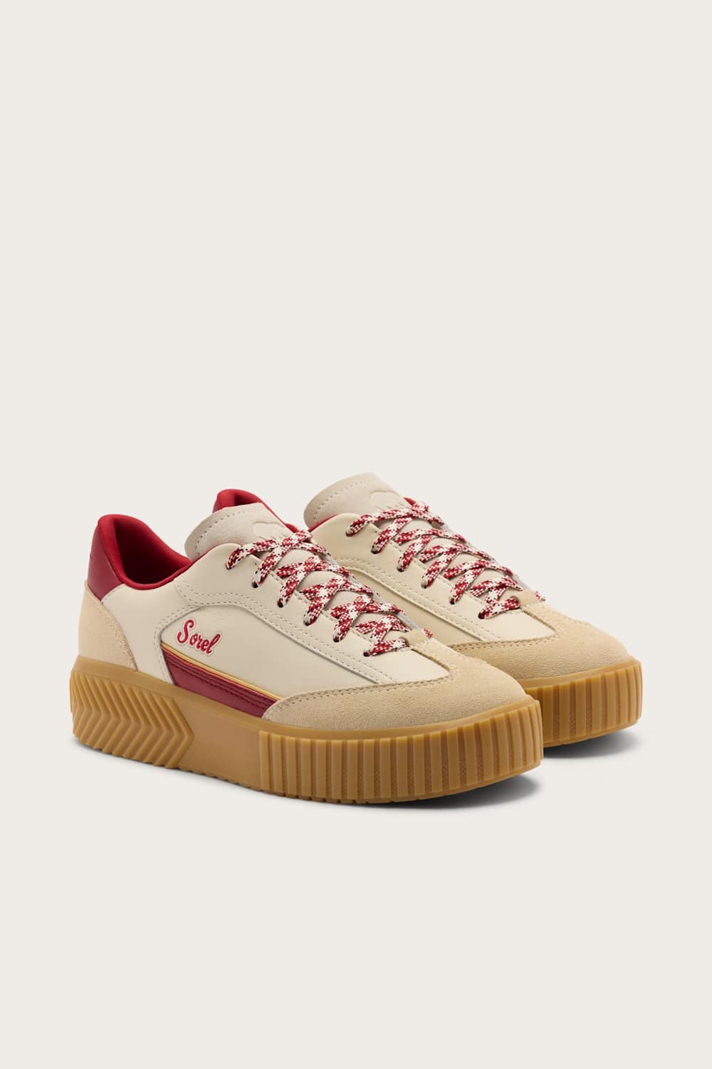 A pair of low-top platform sneakers in beige suede and leather, featuring red patterned laces, a red heel tab, and a tan gum sole, shown against a light beige background.
