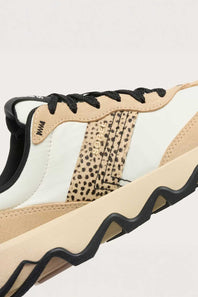 A close-up side view of a low-top sneaker in beige, white, and black, featuring a leopard-print side panel, black laces, and a chunky wavy black sole.