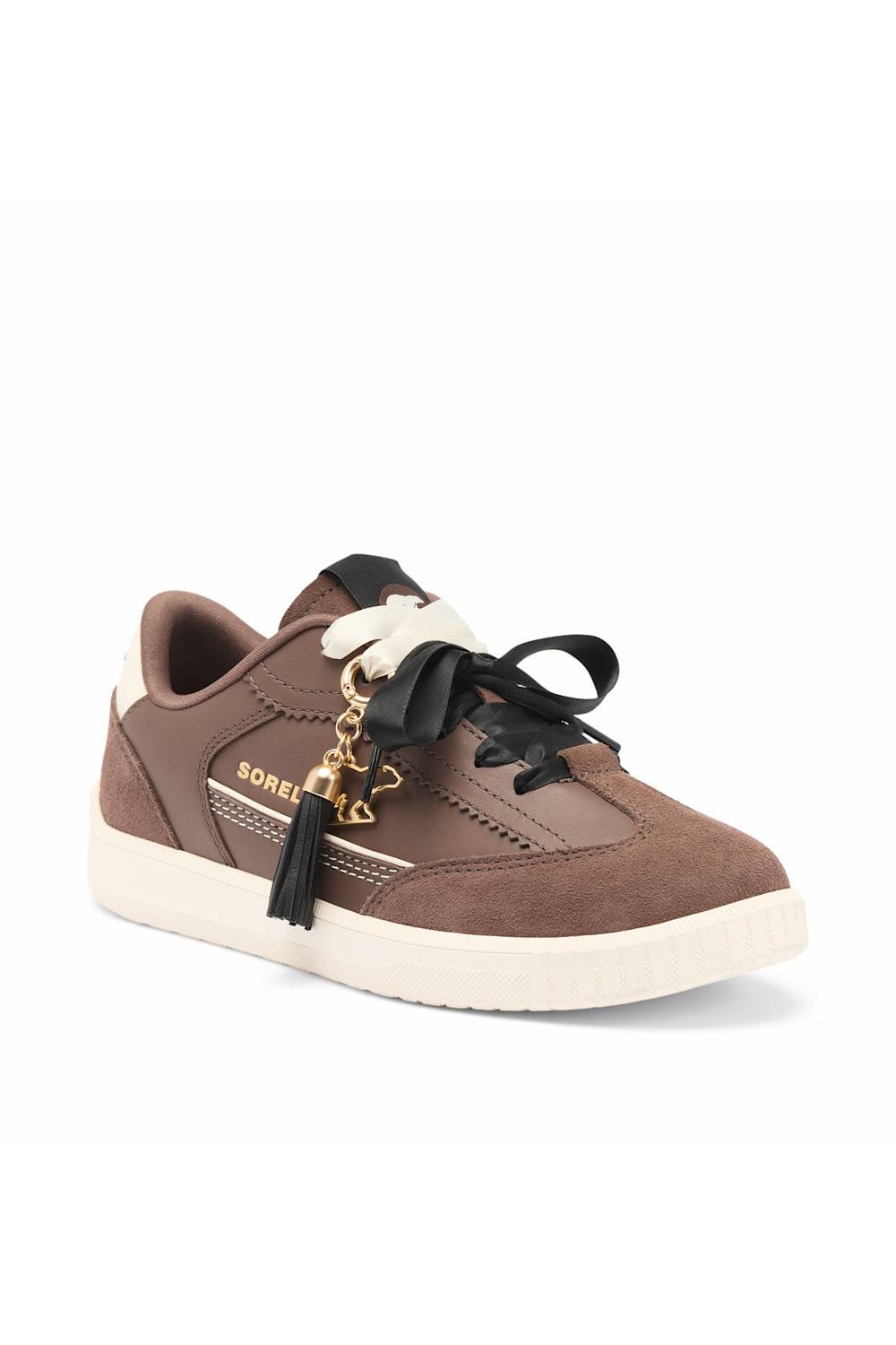 A brown suede and leather sneaker with black laces, a black tassel charm, gold-tone hardware, and a thick off-white sole. 