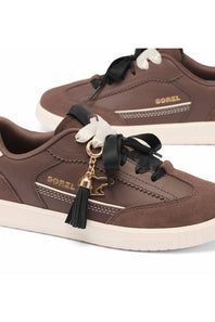 A pair of Sorel brand low-top sneakers in brown suede and leather, featuring black ribbon laces with white bow details, a black tassel charm with a gold horse-shaped keyring, and a cream-colored sole.