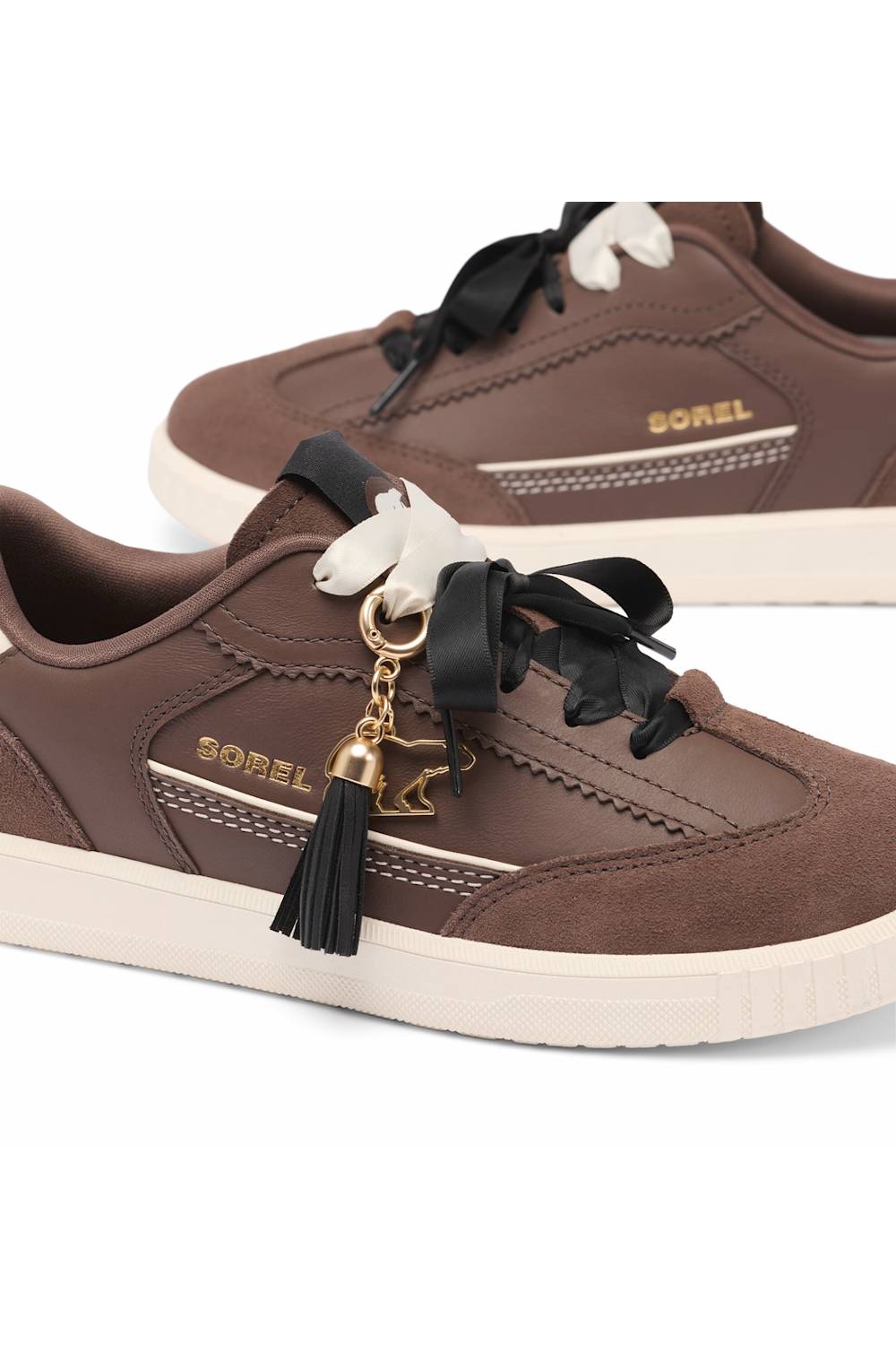 A pair of Sorel brand low-top sneakers in brown suede and leather, featuring black ribbon laces with white bow details, a black tassel charm with a gold horse-shaped keyring, and a cream-colored sole.