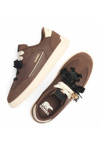 A pair of brown suede sneakers with cream-colored soles, black decorative ribbon laces, and a small gold charm, shown from a top-down view on a white background.