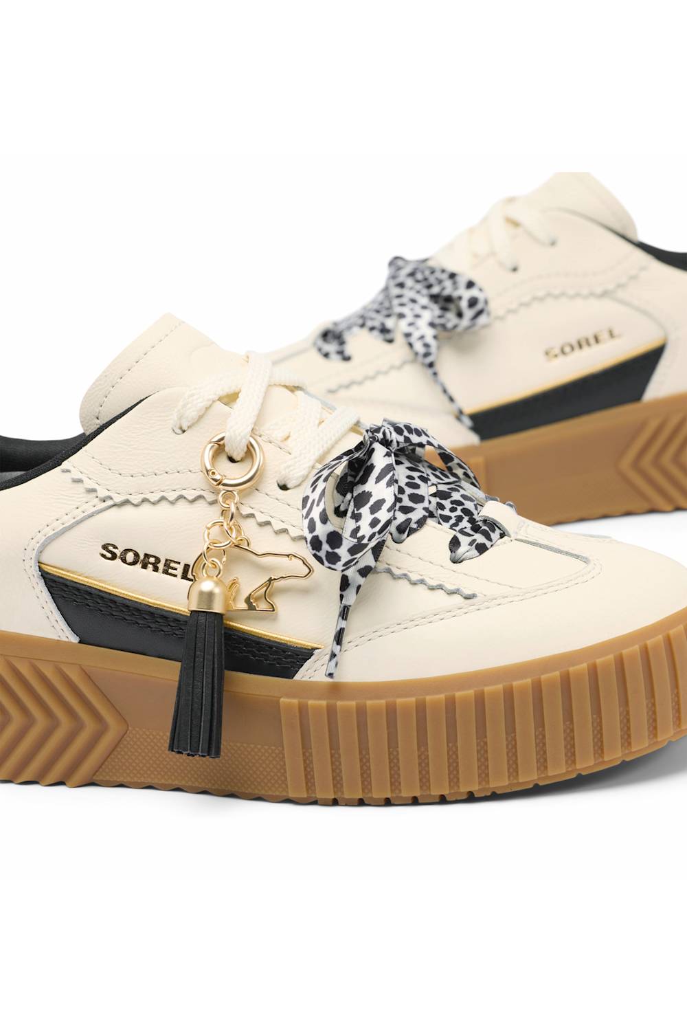 A pair of Sorel platform sneakers in cream and black, featuring brown gum soles, leopard-print ribbon laces, and a black tassel charm with a gold bear logo.