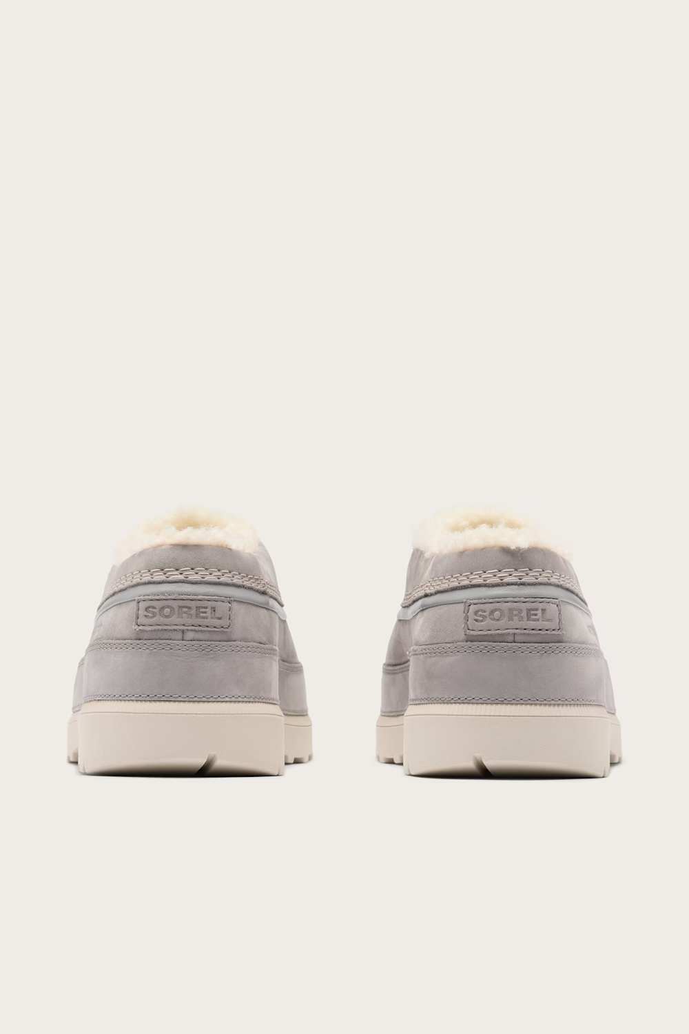 A pair of light gray suede slip-on clogs with white shearling lining, shown from the back on a light cream background. The brand name SOREL is visible on the heel tab.