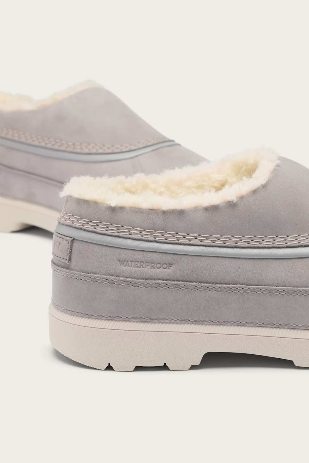 A close-up of a light gray slip-on clogs with a cream-colored shearling lining and a thick off-white sole, featuring a stitched pattern and the word WATERPROOF on the heel.