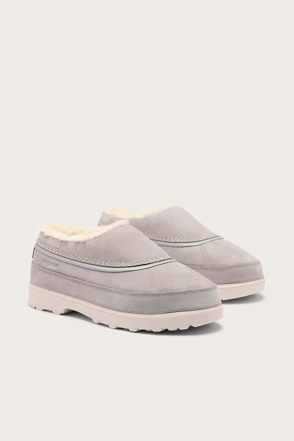 A pair of light gray slip-on clogs with a soft cream-colored shearling lining and a thick, low-profile sole, shown against a solid off-white background.