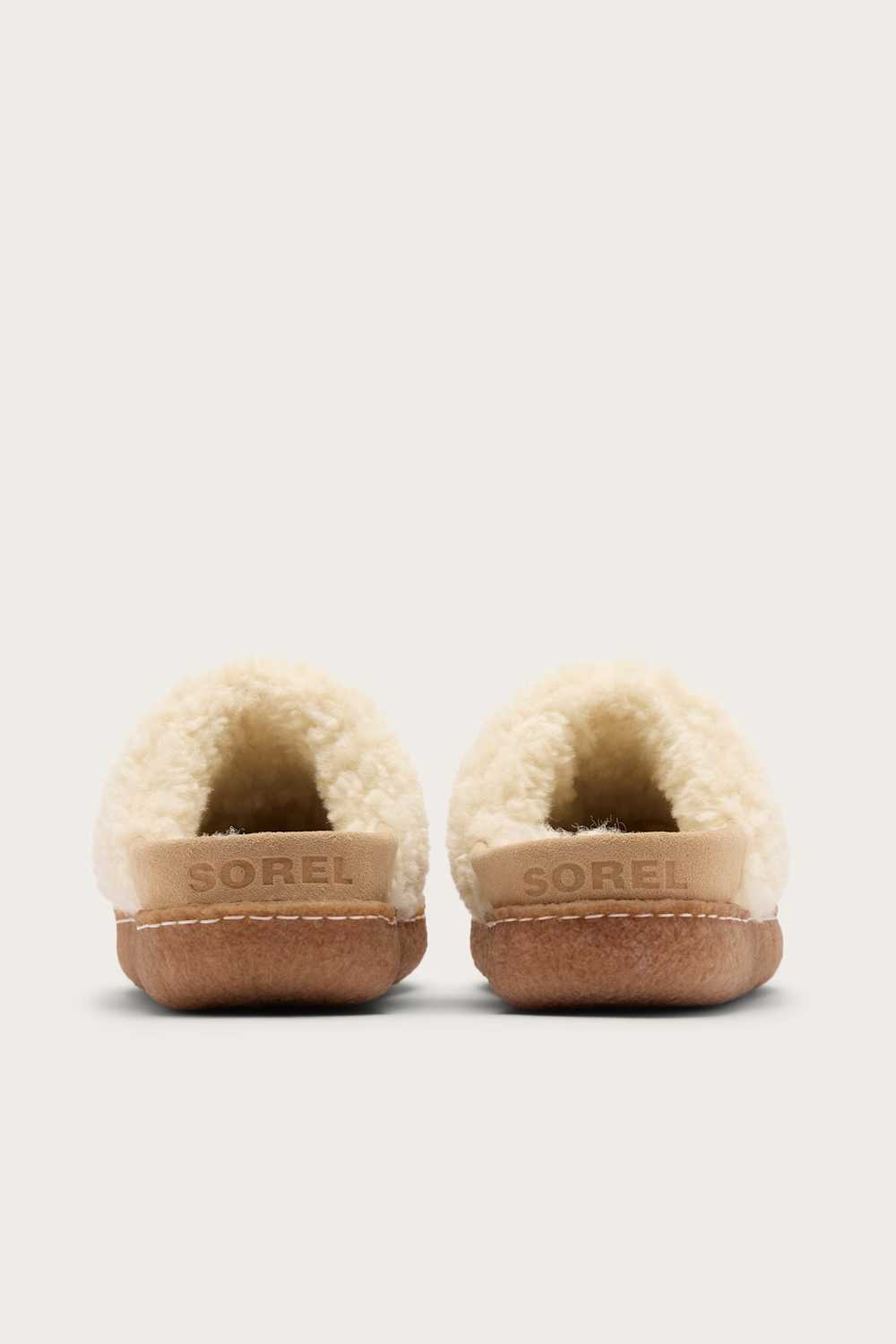 A pair of Sorel slippers shown from the back, featuring a cream-colored shearling lining, a tan leather midsole with the SOREL brand name embossed, and a brown rubber outsole, set against a solid light gray background.