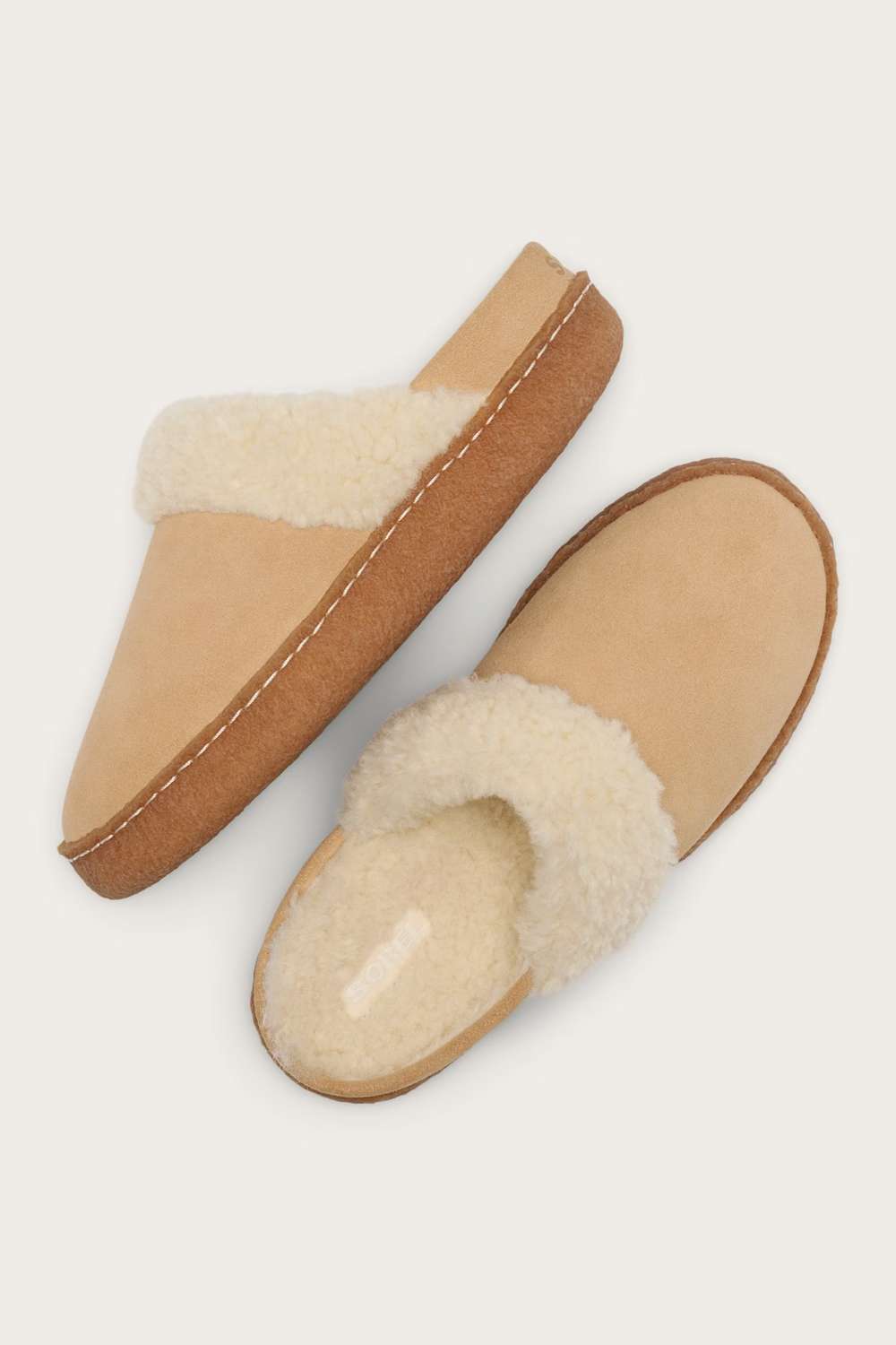 A pair of slip-on mule slippers with beige suede-like uppers, cream shearling-style footbeds and trim, and tan soles, shown on a light beige background.