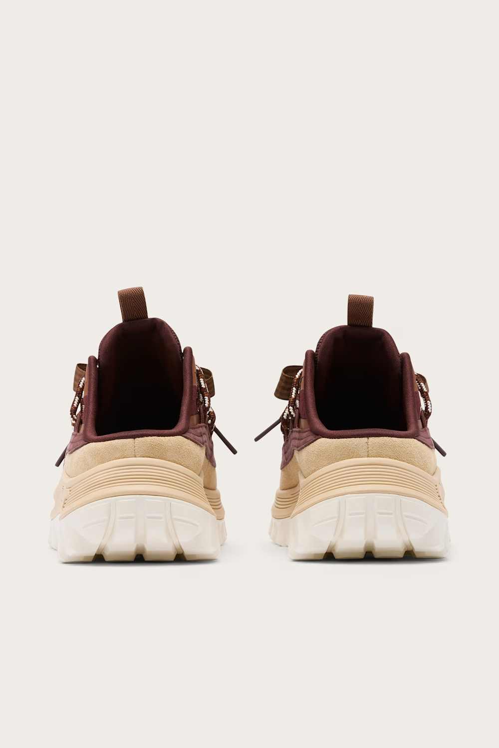 A pair of chunky low-top sneakers shown from the back, featuring a beige and cream textured upper with dark burgundy lining and pull tabs, set against a solid light gray background.