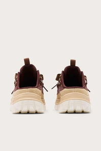 A pair of chunky low-top sneakers shown from the back, featuring a beige and cream textured upper with dark burgundy lining and pull tabs, set against a solid light gray background.