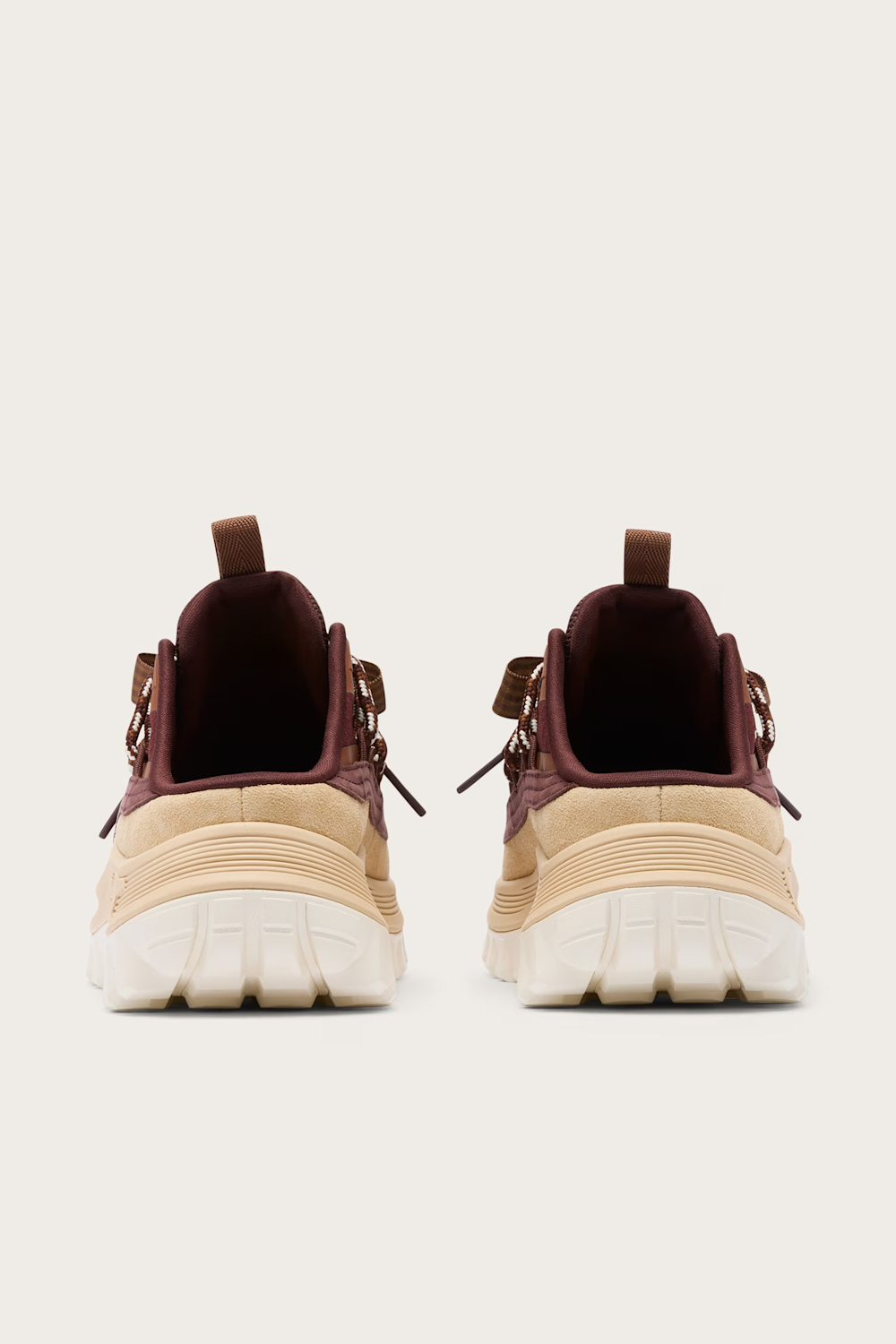 A pair of chunky platform sneakers shown from the back, featuring beige and cream platform soles with dark burgundy and brown upper panels and laces, set against a solid off-white background.