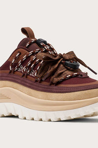 A close-up side view of a Sorel sneaker in dark brown and burgundy with patterned laces, a beige midsole, and a white outsole, set against a light beige background.