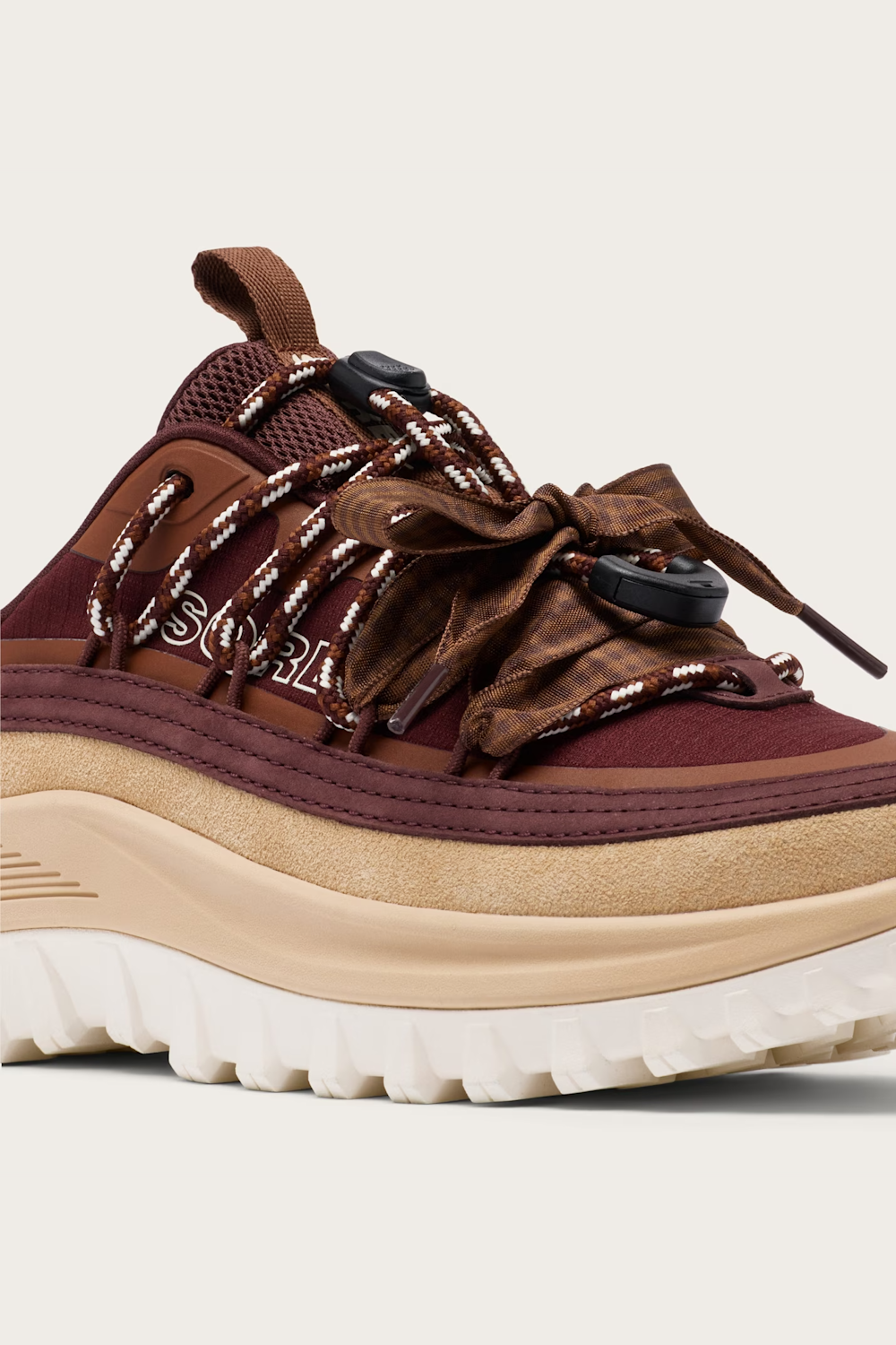 A close-up of a Sorel sneaker in brown and burgundy with patterned laces, a beige midsole, and a white outsole, shown against a light beige background.